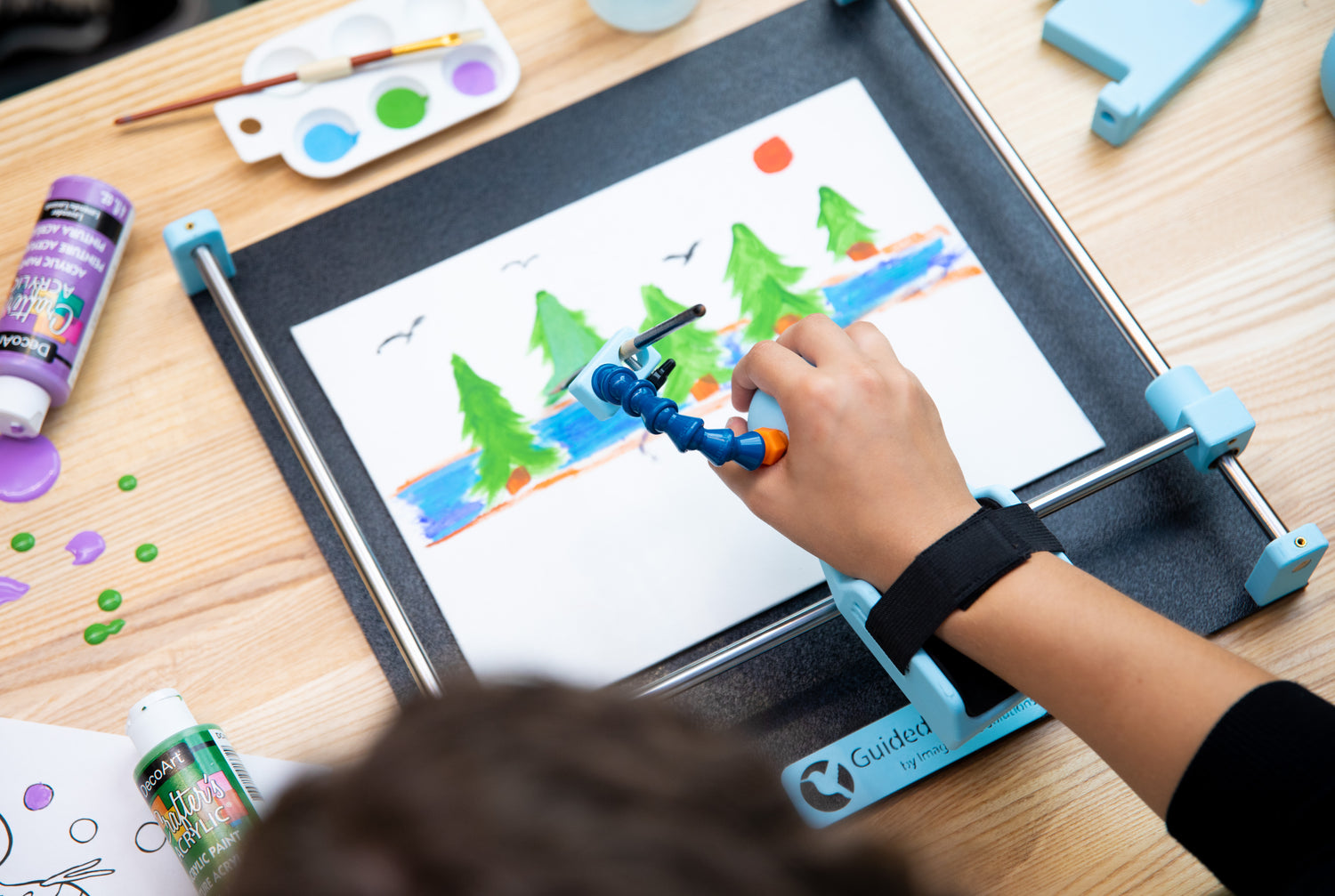 Guided Hands being used to paint a scene of trees beside a river with birds flying overhead